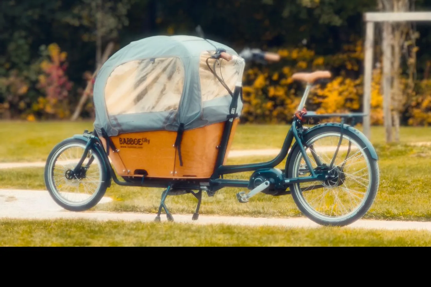 Front-box electric cargo bike with a rain canopy over the child box, parked in a park