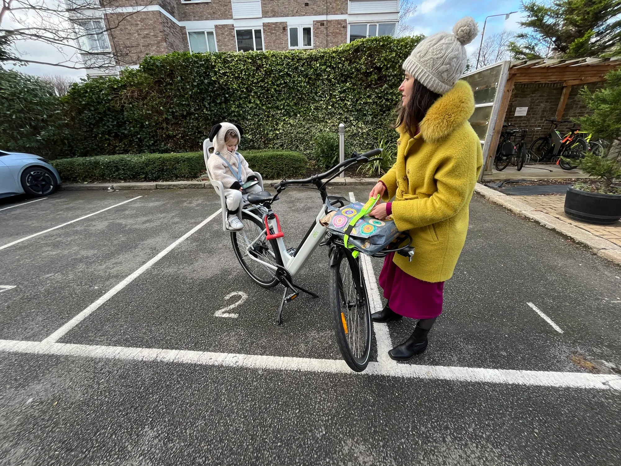 Mum with her toddler in the child seat of a CYCL compact e-bike beside a communal bike shed